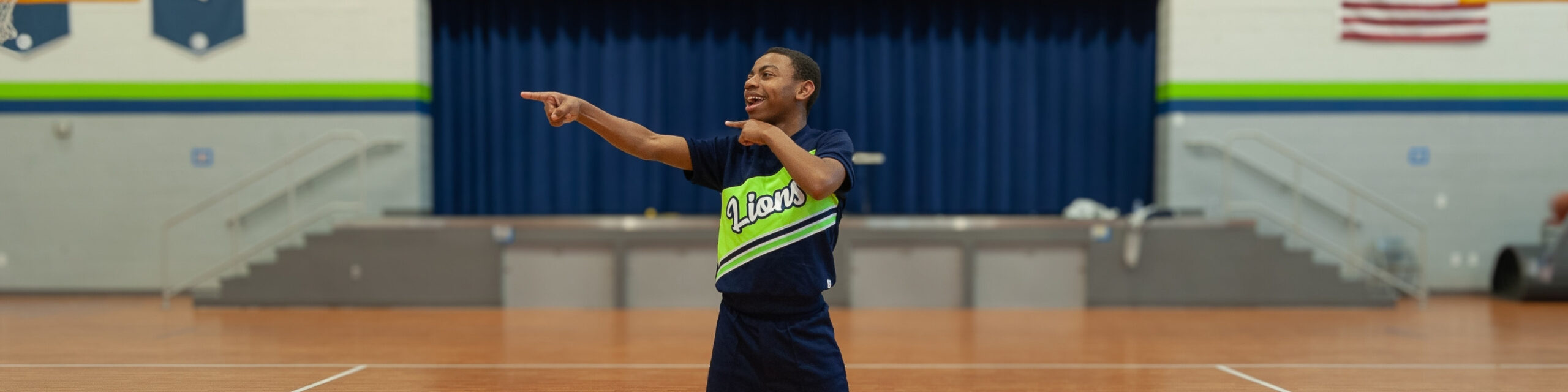 Cheerleader standing in the gym pointing, smiling, and looking to the side.