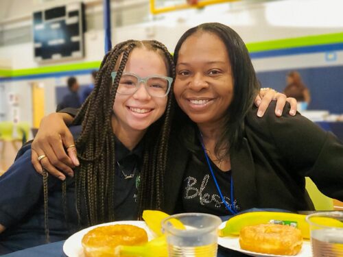 Middle school student sitting around a table arm in arm with her mother, both smiling.