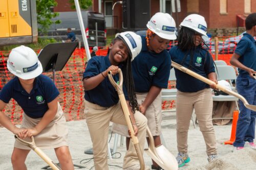 Lower school students wearing hard hats and digging with shovels during the new Lower School building groundbreaking ceremony.